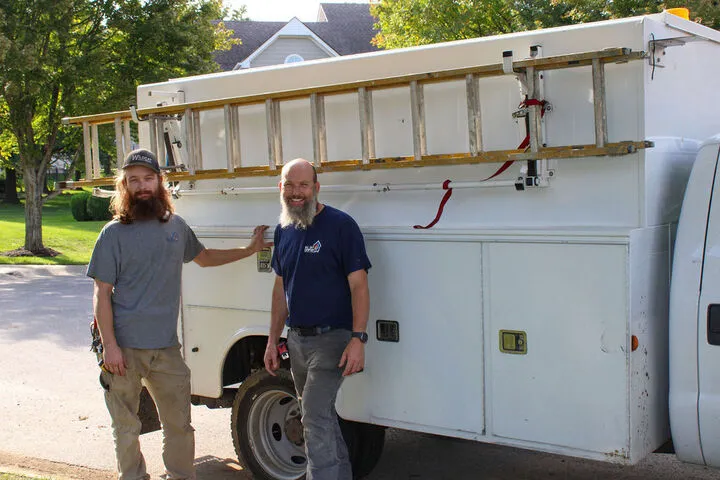 Two All Day Comfort technicians standing next to a service vehicle with a ladder.